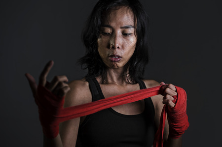 portrait of sporty and fit fighter Asian Chinese woman using wrist wraps wrapping before mma fight sport or boxing workout looking dangerous in badass pose isolated on dark grungeの写真素材