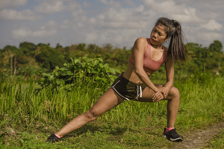 fit and sporty runner Asian woman stretching leg and body after running workout on green field...