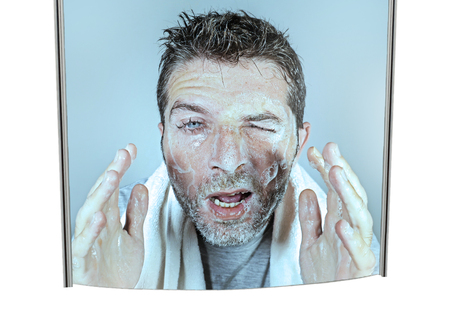 mirror reflection portrait of young attractive and funny man washing face with soap with itchy eyes and messy face expression applying beauty facial skin care treatment in lifestyle conceptの写真素材