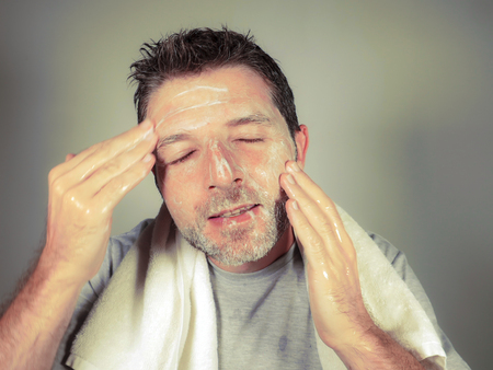 young happy and attractive man applying moisturizer lotion or anti aging beauty cream smiling confident and conscious looking at bathroom mirror in skin care treatment and healthy lifestyleの写真素材