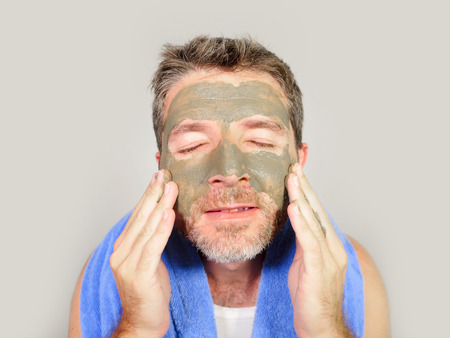 lifestyle isolated portrait of young attractive and happy man with bathroom towel  with green cream on his face applying facial mask in skin care beauty and male cosmetics use conceptの写真素材