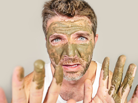 lifestyle funny portrait of young attractive and surprised man horrified looking himself on bathroom mirror ugly and weird applying green cream on his face in facial mask male beauty product conceptの写真素材