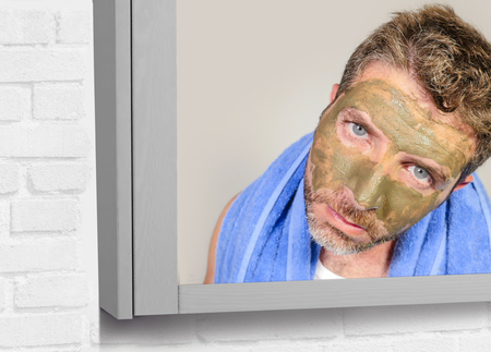 lifestyle funny portrait of young attractive and surprised man horrified looking himself on bathroom mirror ugly and weird applying green cream on his face in facial mask male beauty product conceptの写真素材