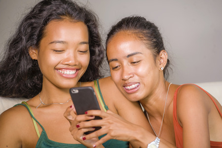 young beautiful and happy Asian girlfriends couple or sisters enjoying internet social media having fun together with mobile phone at  home sofa couch smiling playful and laughing in girls friendshipの写真素材
