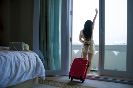 lifestyle portrait of young happy and beautiful Asian Korean tourist woman arriving to luxury hotel room opening balcony windows staring around delighted and excited with suitcaseの写真素材