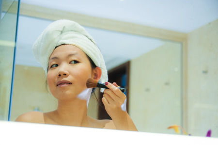 lifestyle fresh portrait of young happy and beautiful Asian Korean woman at home or hotel bathroom wrapped in toilet towel applying makeup cheerful and natural in female beauty and skin careの写真素材