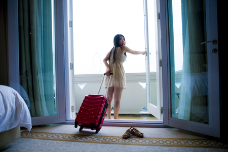 lifestyle portrait of young happy and beautiful Asian Chinese tourist woman arriving to luxury hotel room opening balcony windows staring around delighted and excited with suitcaseの写真素材