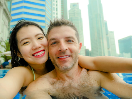young happy and attractive playful couple taking selfie picture together with mobile phone at luxury urban hotel infinity pool enjoying holidays honeymoon travel in diversity ethnicity and loveの写真素材