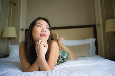 lifestyle indoors portrait of young beautiful and happy Asian Korean woman lying on bed in the morning at home bedroom or hotel room feeling cheerful and relaxed smiling playfulの写真素材