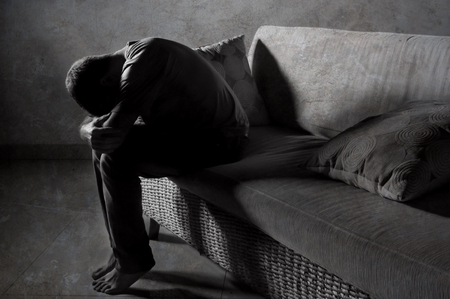 lifestyle dramatic light portrait of young sad and depressed man sitting at shady home couch in pain and depression feeling stressed and desperate crying alone suffering anxiety crisisの写真素材