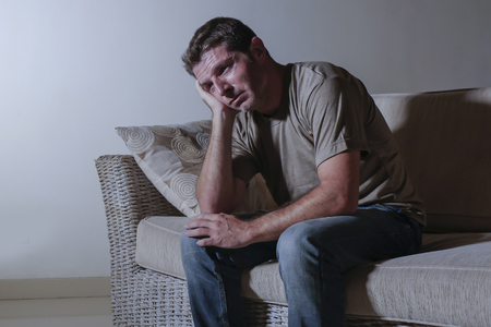 lifestyle dramatic light portrait of young sad and depressed man sitting at shady home couch in pain and depression feeling stressed and desperate crying alone suffering anxiety crisisの写真素材