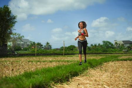 healthy lifestyle portrait of young happy and fit Southeast Asian Thai runner woman in running workout outdoors at green field background in body exercise health care and fitness conceptの写真素材