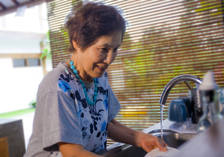 lifestyle portrait of senior happy and sweet Asian Japanese retired, woman cooking at home kitchen alone neat and tidy washing the dishes smiling cheerful in house chores conceptの写真素材