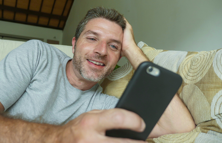 lifestyle indoors portrait of young happy and attractive man at home sofa couch using internet social media app on mobile phone networking relaxed and cheerful from modern apartmentの写真素材
