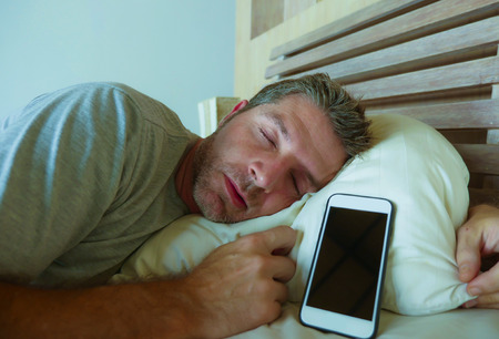 lifestyle indoors portrait of young internet addict man sleeping on bed holding mobile phone in his...
