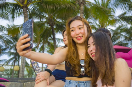 lifestyle portrait of group with young happy and attractive Asian Chinese and Korean women hanging out , girlfriends enjoying holidays trip in tropical resort taking selfie picture with mobile phoneの写真素材