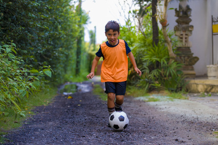8 or 9 years old happy and excited kid playing football outdoors in garden wearing training vest running and kicking soccer ball , the kid having fun practicing sportの写真素材