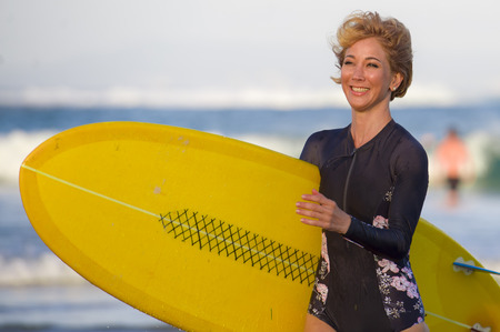 young attractive and happy blonde surfer girl in beautiful beach carrying yellow surf board walking out of the water enjoying summer holidays at tropical island in in woman surfing lifestyle conceptの写真素材