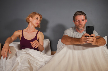 husband or boyfriend using mobile phone in bed and suspicious frustrated wife or girlfriend feeling...