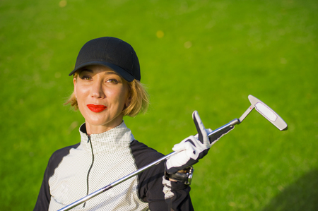 lifestyle outdoors portrait of young beautiful and happy woman at playing golf holding ball and putter club smiling cheerful in stylish golfing clothes isolated on green put backgroundの写真素材