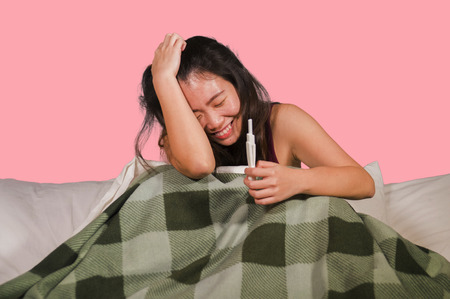 young happy and excited Asian Chinese woman in bed holding pregnancy test checking surprised positive pregnant result smiling ecstatic and joyful expecting baby elated feeling  blessedの写真素材