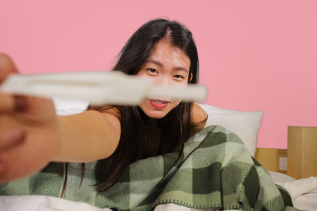 young happy and excited Asian Korean woman in bed holding pregnancy test checking surprised positive...