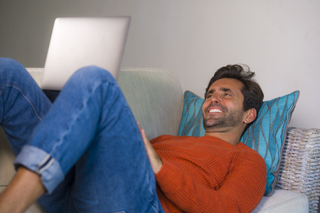 young happy and attractive man working relaxed with laptop computer at modern apartment living room lying at sofa couch typing and networking in entrepreneur success and lifestyle conceptの写真素材