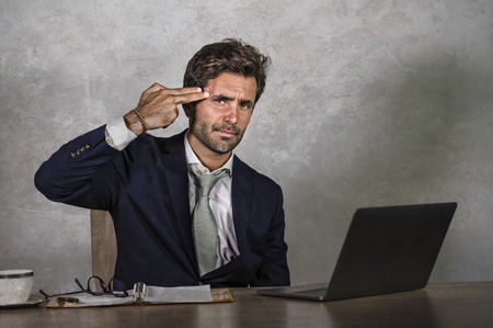 young sad and depressed business man working overwhelmed and frustrated on laptop computer office desk feeling upset and stressed pointing hand gun fingers to his head as suicide gestureの写真素材