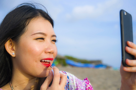 young happy and beautiful Asian Chinese woman retouching her makeup applying red lipstick on her lips using mobile phone as mirror at tropical paradise beach during summer holidays travelの写真素材
