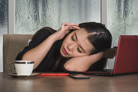 young beautiful sad and depressed Asian Japanese business woman working exhausted and frustrated at office computer desk with rain on the window suffering depression and anxiety problemの写真素材