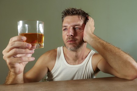 young wasted and depressed alcohol addict man in dirty singlet drinking glass of whiskey feeling desperate suffering alcoholism problem and booze addiction on isolated background in alcoholic conceptの写真素材