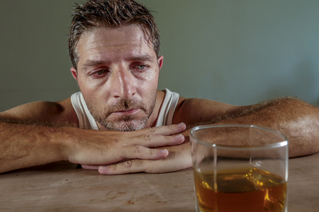 young wasted and depressed alcohol addict man in dirty singlet drinking glass of whiskey feeling desperate suffering alcoholism problem and booze addiction on isolated background in alcoholic conceptの写真素材
