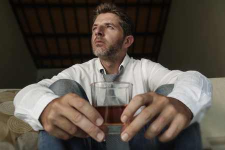 young attractive sad and desperate drunk addict businessman at home sofa couch drinking holding whiskey glass feeling depressed in alcohol addiction alcoholism problem and depressionの写真素材