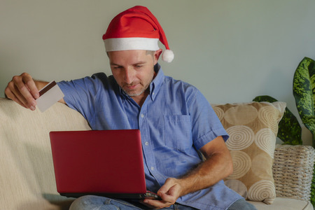 young attractive and happy man in Santa Klaus Christmas hat using laptop computer for buying xmas presents and gifts online with credit card in internet banking and electronic commerce and businessの写真素材