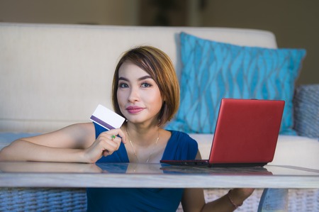 young beautiful and happy woman at home living room holding credit card using laptop computer for banking and online shopping smiling relaxed in e-business and internet commerce conceptの写真素材