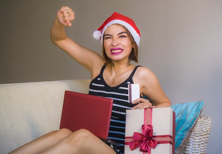 young happy and attractive woman relaxed at apartment couch in Christmas hat using laptop computer...