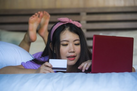 young beautiful and happy Asian Korean woman using credit card internet banking on laptop computer at home in bed smiling cheerful shopping online buying in e-commerce and lifestyle conceptの写真素材