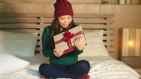 young happy and beautiful Asian Chinese woman in winter hat holding Christmas giftbox with ribbon smiling excited and cheerful receiving x-mas present sitting at home bed opening boxの写真素材