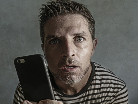 young weird and crazy mobile phone addict man using cell compulsively with weird and freak face expression in internet social media addiction and obsession isolated on even backgroundの写真素材