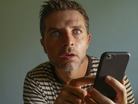 young weird and crazy mobile phone addict man using cell compulsively with weird and freak face expression in internet social media addiction and obsession isolated on even backgroundの写真素材