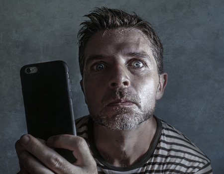 young weird and crazy mobile phone addict man using cell compulsively with weird and freak face expression in internet social media addiction and obsession isolated on even backgroundの写真素材