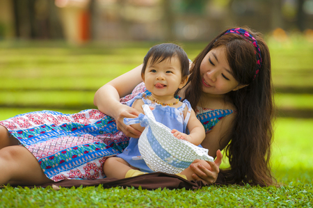 happy and playful Asian Korean woman as loving mother enjoying with sweet and beautiful daughter baby girl sitting together playing on grass at city park in motherhood responsibility and loveの写真素材