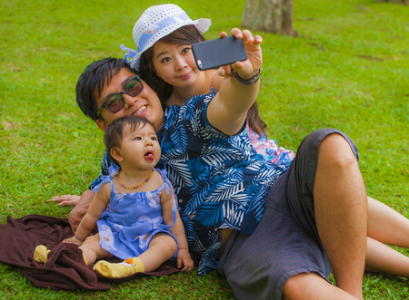 young happy and loving Asian Japanese family with parents and sweet baby daughter at city park together with father taking selfie pic with mobile phone for uploading on internet social mediaの写真素材