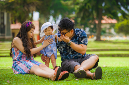 young happy and loving Asian Chinese parents couple enjoying together with sweet daughter baby girl sitting on grass at green city park in healthy beautiful parenthood and childhood conceptの写真素材