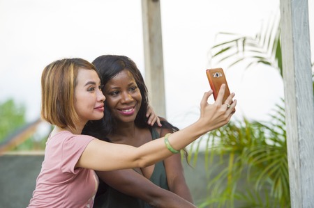 young happy and beautiful black afro American woman enjoying holidays at tropical resort with Asian girlfriend taking selfie picture with mobile phone together in women diversity race friendshipの写真素材