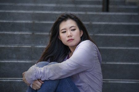 young sad and depressed Asian Japanese student woman or bullied teenager sitting outdoors on street...
