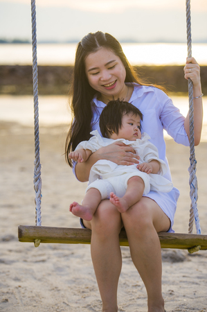 lifestyle outdoors portrait of young sweet and happy Asian Chinese woman holding baby girl swinging together at beach swing on Summer sunset in mother and little daughter love conceptの写真素材