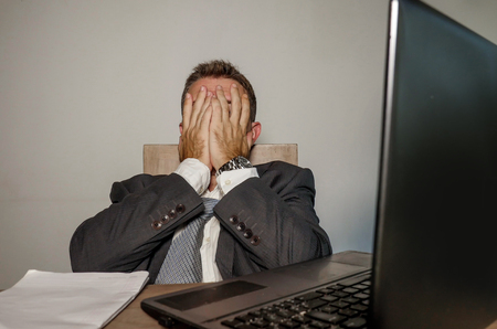 young desperate and depressed business man suffering problem and depression at office desk working with laptop computer feeling frustrated and overwhelmed on isolated backgroundの写真素材