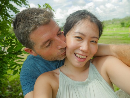young beautiful and happy mixed ethnicity couple beautiful Asian Chinese woman and white man in love taking selfie picture outdoors enjoying romantic holidays trip in tropical honeymoon vacationの写真素材
