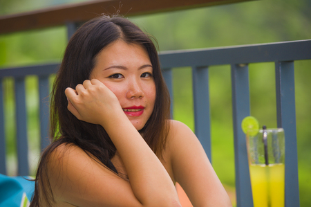 young happy and beautiful Asian Chinese tourist woman sitting relaxed outdoors at snack cafe drinking orange juice enjoying the view of tropical jungle in holidays travel destination conceptの写真素材